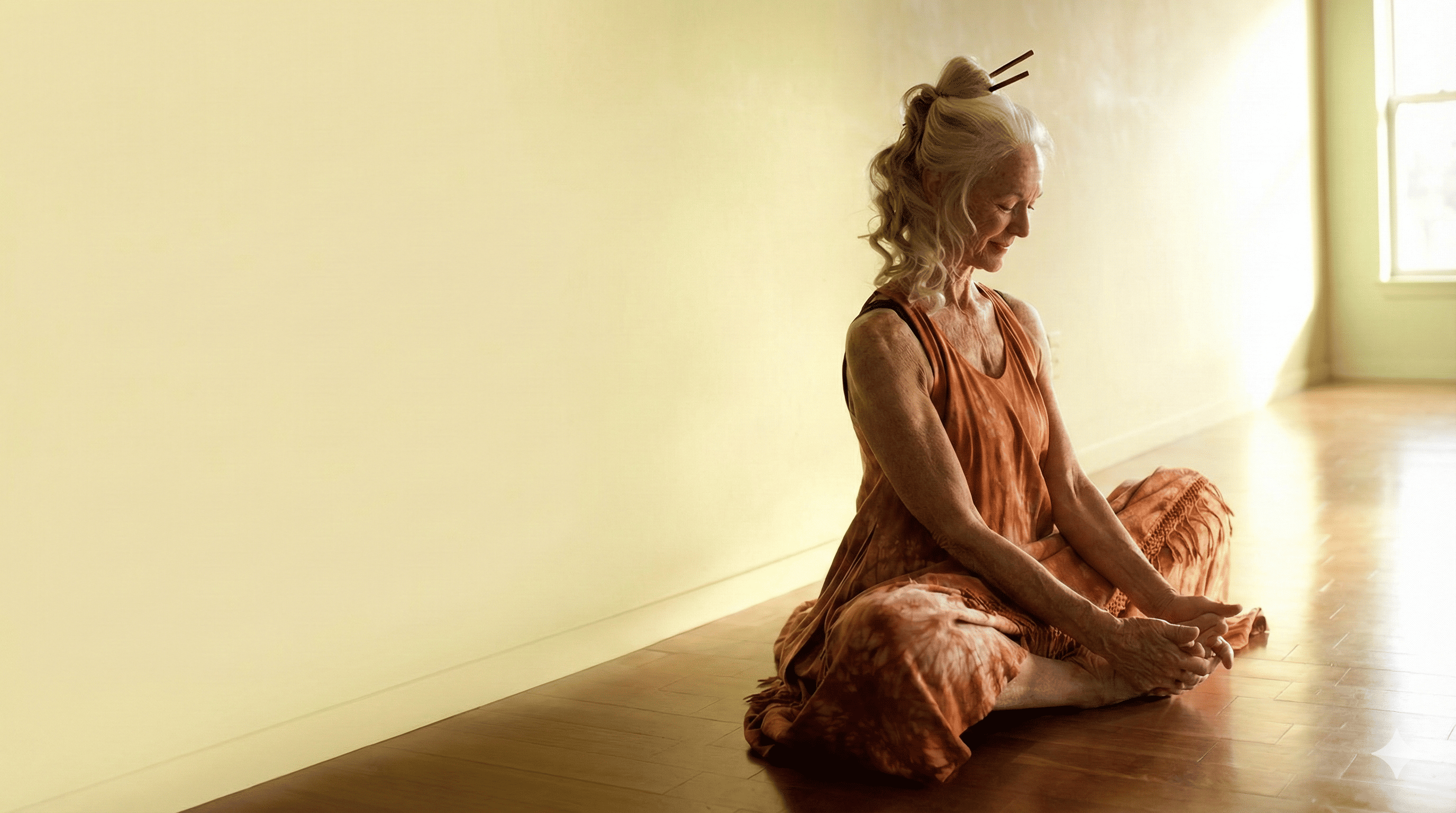 "Serene elderly woman with grey hair tied in a bun, sitting in a cross-legged meditation pose on a wooden floor, bathed in warm natural sunlight, illustrating the anti-aging benefits of Kayakalpa Yoga."