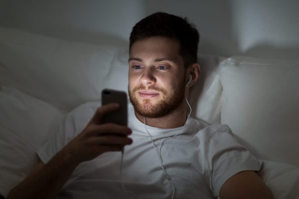 technology, internet, communication and people concept - happy smiling young man with smartphone and earphones listening to music in bed at night